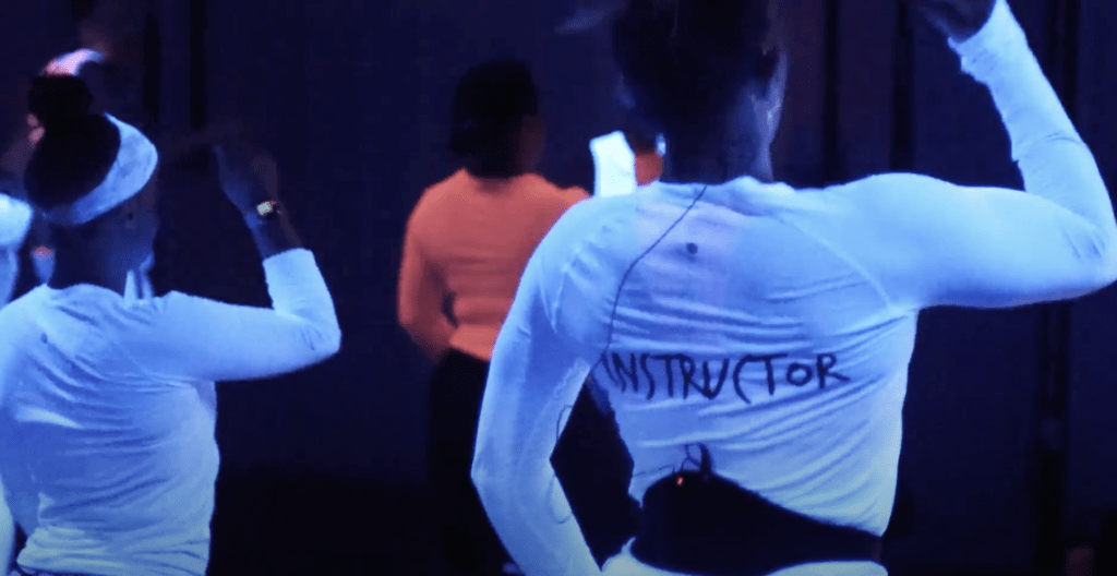 A fitness instructor with the word "INSTRUCTOR" on her back teaches a hip hop cardio class.