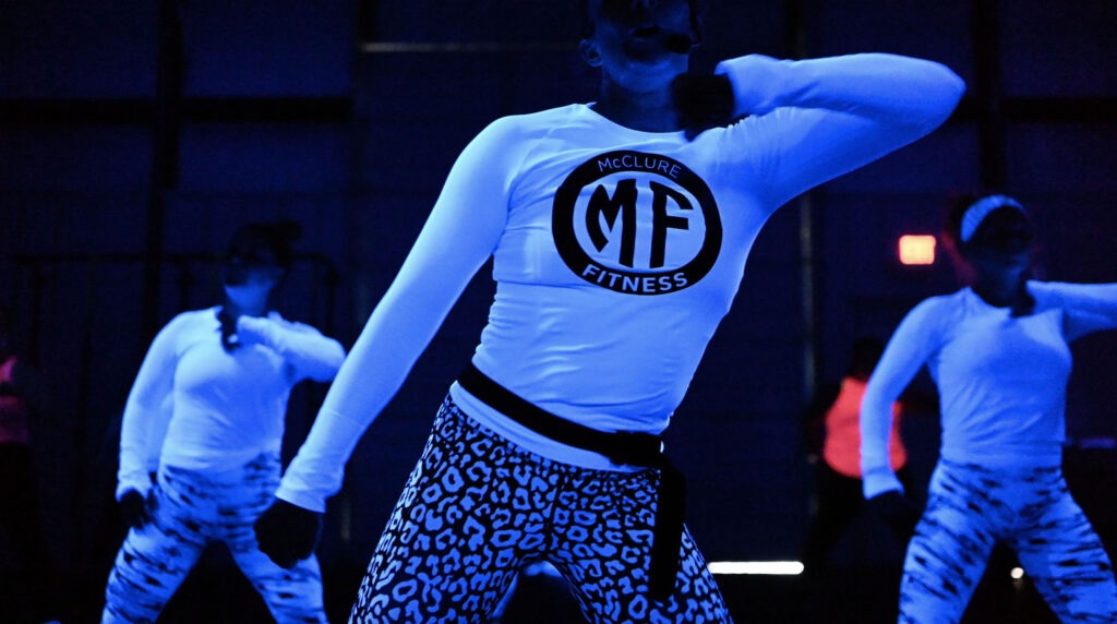 A fitness instructor with the MF logo on her shirt teaches a hip hop cardio class in a dark room with neon clothing.