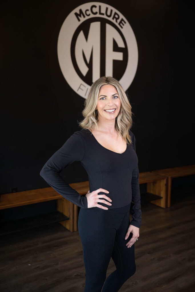 A fitness instructor stands in front of the McClure Fitness logo smiling.